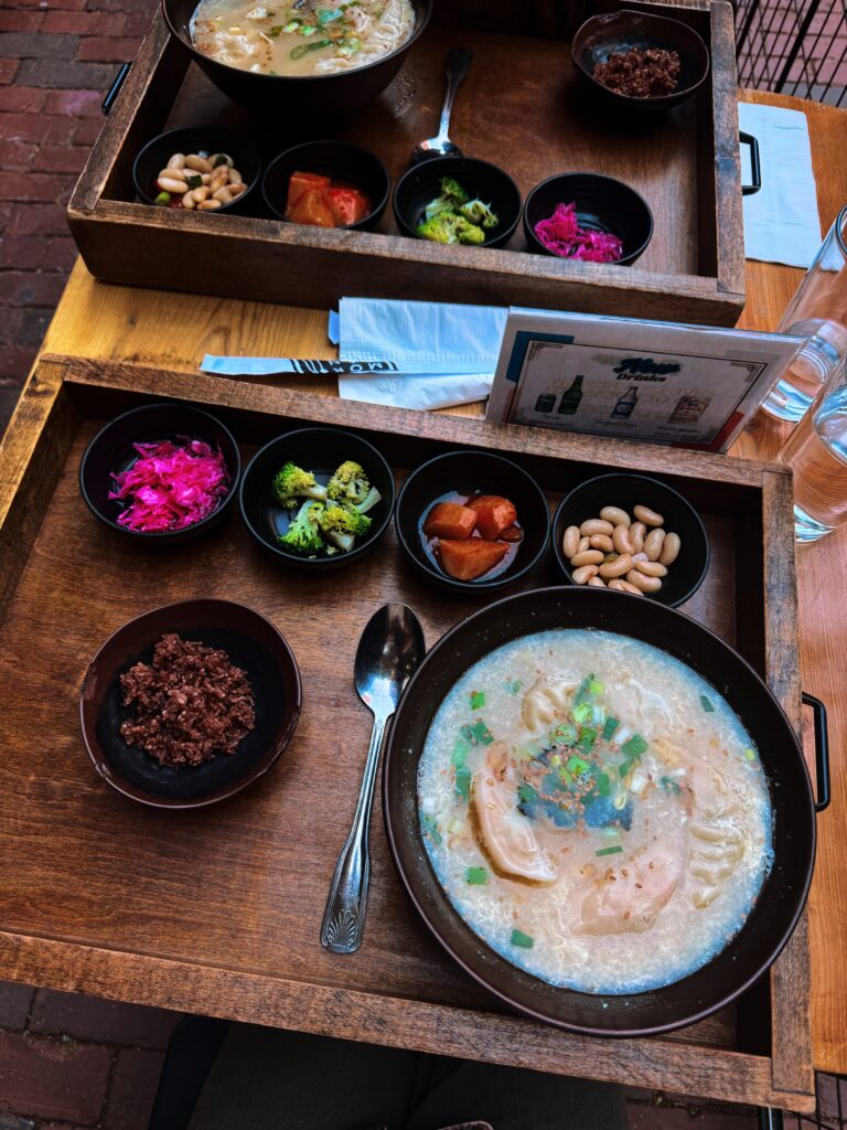 A wooden tray featuring a balanced, whole food meal with a bowl of dumpling soup topped with scallions, served alongside small dishes of pickled cabbage, broccoli, white beans, stewed tomatoes, and a chocolate-like dessert. The meal is photographed outdoors on a wooden table with brick pavement in the background.